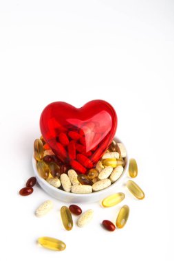 Hap kullanımı. İçinde haplar olan şeffaf kırmızı bir kalp farklı haplarla bir fincan tabağında yatıyor. Beyaz arkaplanda.