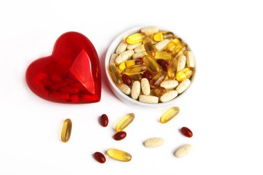 İçinde haplar olan şeffaf kırmızı bir kalp, içi haplarla dolu bir fincan altlığının yanında yatıyor. Beyaz arkaplanda.