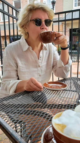 ストリートカフェでサングラスと白いブラウスを着た大人の女性がコーヒーを飲み — ストック写真
