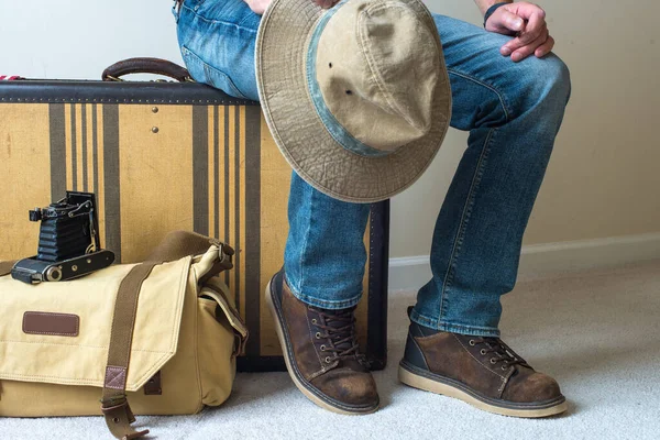 ジーンズと古いブーツを着た男性が出発を待っているヴィンテージのスーツケースに座っています — ストック写真