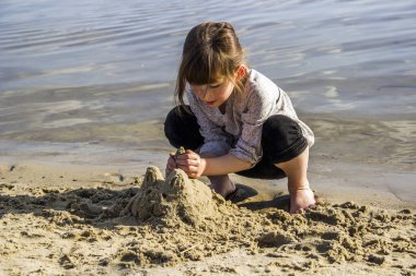 Küçük bir kız nehir sahilinde kedi heykeli yapıyor.