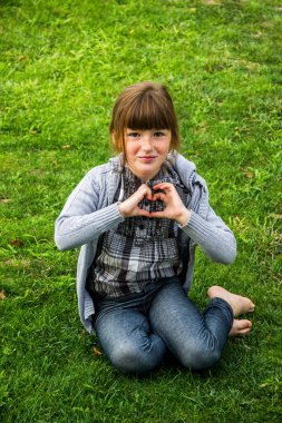 happy little caucassian girl sittig on the green grass and make handly sign of heart