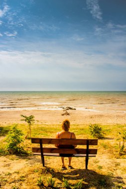 Deniz kıyısının önündeki bankta oturan ve denize bakan bir kadın.