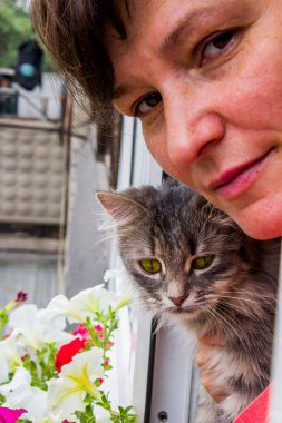 Gülümseyen bir kadın ve balkonda büyük gözleri olan gri bir kedinin yakın çekimi.