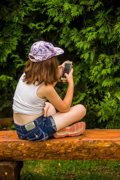 Little Girl Sit Wooden Bench Look Smartphone Thuja Tree — Stockfoto