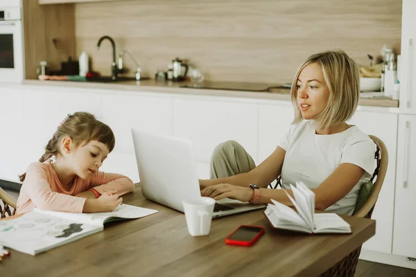 一个女人在家里和她的小女儿一起工作 坐在旁边画画 妈妈坐在厨房里 用笔记本电脑 舒适的办公室工作场所 远程工作概念 与孩子一起在家工作概念 — 图库照片