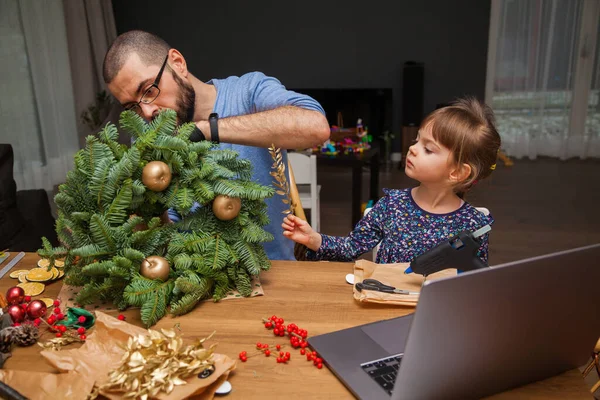 Man and his little daughter learning to decorate a Christmas wreath watching lesson online. Family making Christmas wreath. New year holiday decoration. Christmas tree wreath decoration DIY.
