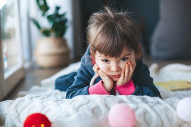 Sıkıcı küçük kız kışın pencerenin yanında yün bir battaniyenin üzerinde yatıyor.