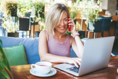 Gülümseyen kadın, 30 küsur yaşında cep telefonuyla konuşuyor, açık bir kafede oturuyor, dizüstü bilgisayar kullanarak çalışıyor. Konforlu ofis işyeri, uzaktan çalışma ve ev konseptinde çalışma.