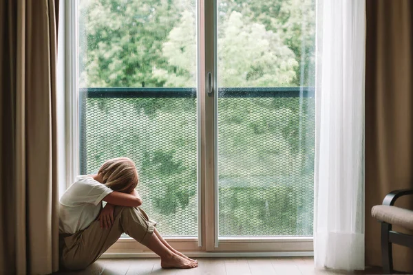 沮丧的女人坐在靠近窗户的地板上抱着膝盖 寂寞的女孩待在家里 年轻而多愁善感的女人在思考问题时感到沮丧 — 图库照片