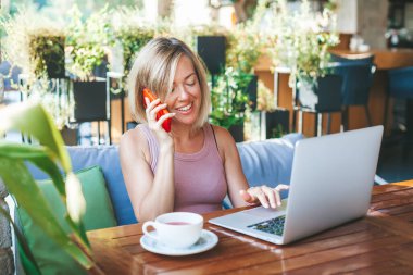 Gülümseyen serbest çalışan kadın cep telefonuyla konuşuyor, güneşli açık kafede oturuyor, dizüstü bilgisayarla çalışıyor. Konforlu ofis işyeri, uzaktan çalışma ve ev konseptinde çalışma.