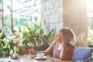 Genç düşünceli kadın SMS mesajları için akıllı telefonu tutuyor ve kullanıyor. Güneşli kafede dokunmatik cep telefonu kullanıyor. Yaşam tarzı, teknoloji ve sosyal medya ağı kavramı.