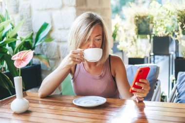 Sarışın kadın çay içiyor ve SMS mesajları için akıllı telefon kullanıyor. Güneşli kafede dokunmatik cep telefonu yazıyor. Yaşam tarzı, teknoloji ve sosyal medya ağı kavramı.