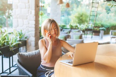 Ciddi kadın serbest çalışan telefonla konuşuyor, güneşli bir kafede oturuyor. Bir dizüstü bilgisayar üzerinde çalışıyor. Konforlu ofis işyeri, uzaktan çalışma ve ev konseptinde çalışma.
