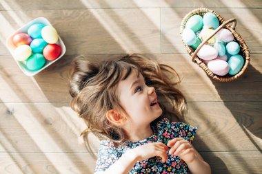 Neşeli gülümseyen küçük kız pastel renkli Paskalya yumurtalarını sepete koyup yerde yatıyor.