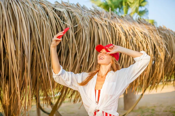 站在海滩上 戴着红帽子 面带微笑的漂亮女人在电话里自作主张 — 图库照片