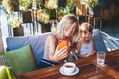 Sarışın kadın ve küçük kızı kafede görüntülü sohbet için akıllı telefon kullanıyorlar. Yaşam tarzı, teknoloji ve iletişim konsepti.