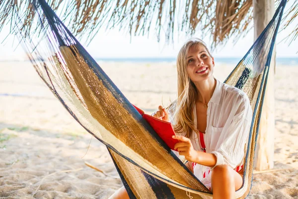 美丽的微笑的女人坐在海滩上的吊床上 用红色的规划器写下了一些东西 舒适环境中的生活或工作规划 — 图库照片