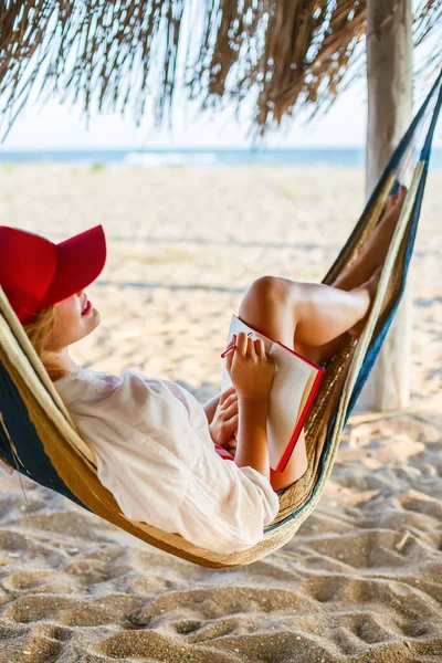 Kobieta Czerwonej Czapce Pisze Coś Czerwonym Planerze Leżącym Hamaku Plaży — Zdjęcie stockowe