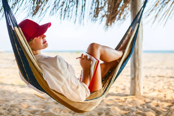 Woman in red cap writing something in her red planner lying in a hammock on a beach. Life or job planning in comfortable environment.