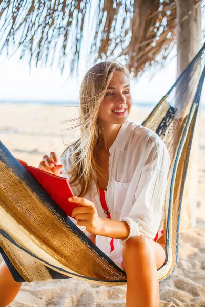 美しいブロンドの女性はビーチでハンモックに座って赤いプランナーに何かを書いています 快適な環境での生活や仕事の計画 — ストック写真