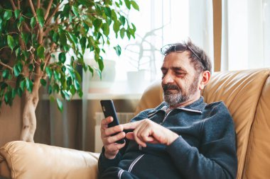 Olgun bir adam kanepeye oturmuş, mesajlaşıyor, arıyor ya da modern bir akıllı telefondan kitap okuyor. Teknoloji kavramını kullanan yaşlılar.