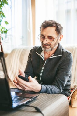60-65 yaşlarında, gözlüklü, dizüstü bilgisayarının önünde oturan ve akıllı telefon kullanan olgun bir adam. Teknoloji kavramını kullanan yaşlılar.