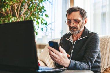 60-65 yaşlarında, gözlüklü bir adam dizüstü bilgisayarın önündeki koltukta oturuyor ve modern bir akıllı telefon kullanıyor. Teknoloji kavramını kullanan yaşlılar.
