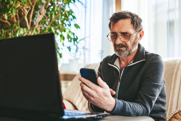 60-65 yaşlarında, gözlüklü bir adam dizüstü bilgisayarın önündeki koltukta oturuyor ve modern bir akıllı telefon kullanıyor. Teknoloji kavramını kullanan yaşlılar.