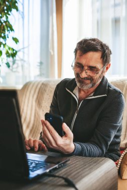 60-65 yaşlarındaki gülümseyen olgun bir adam bir kanepede oturuyor ve modern bir akıllı telefon kullanıyor. Teknoloji kavramını kullanan yaşlılar.