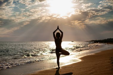 Sahilde ağaç pozisyonunda duran bir kadının arka görüntüsü. Güneşi seyrediyor. İç uyum ve vücut bakımı.