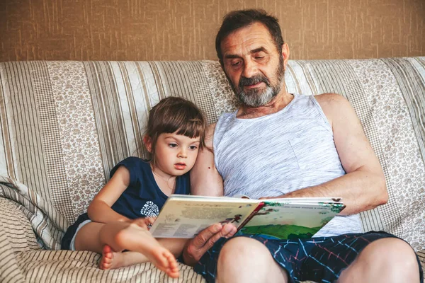 Nagyapa Könyvet Olvas Unokájának Egy Kanapén — Stock Fotó