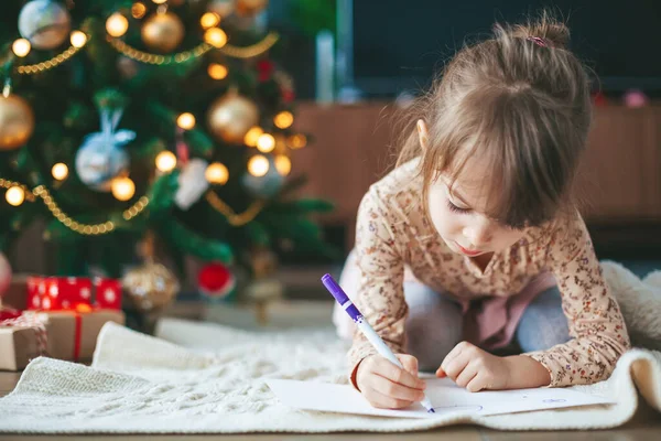 Cute little girl writing letter to Santa Claus or writing dreams of a gift with a Christmas tree on a background. Merry Christmas and Happy Holidays!