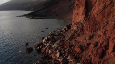El Hierro adasının batımında, volkanik dağların ve beyaz bulutların zeminine serpilmiş, çökmüş bir kaya parçalarıyla, parlak kırmızı paslı uçurumun kenarına doğru eğik duran hava aracı.