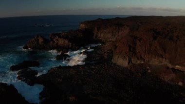 Kahverengi sert lavın kayalık sahil şeridi ve okyanus dalgası dalgaları geçen günün son ışınlarında El Hierro, Kanarya Adaları 'ndaki El Hierro adasının Verodal plajı yakınlarında dalgalanır.