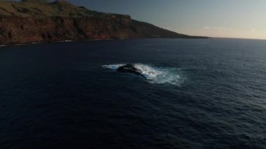 Atlantik Okyanusu dalgaları tarafından sürüklenen düz kaya parçasının etrafında uçan yay, El Hierro ve Verodal adalarının volkanik manzarasının arka planına karşı çekilmiştir.