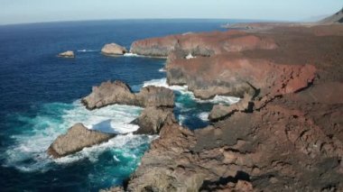 Kara kahverengi sert lavlardan oluşan kayalık bir sahil kulesi ve Kanarya Adaları 'ndaki El Hierro adasının Verodal plajı yakınlarındaki güneşli bulutsuz havada okyanus dalgası sörfü.