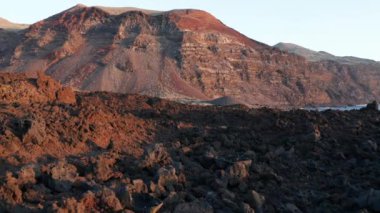 El Hierro adasının kızıl küllü dağının arka planında uzun gölgeli batan güneşin ışığında, siyah kahverengi, katı volkanik lav arazisinin üzerinde yavaşça uçan kule.