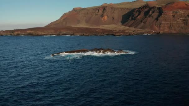 晴れた日にエルヒエロ島の火山の風景を背景に大西洋の波によって洗われた平らな岩の周りに空中アークショット — ストック動画