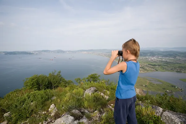 Boy looking through binoculars from the top of the mountain to t — Stock Photo, Image