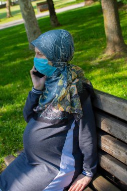 Yüz maskesi takmış, akıllı telefondan konuşan Müslüman hamile kadının profil görüntüsü.