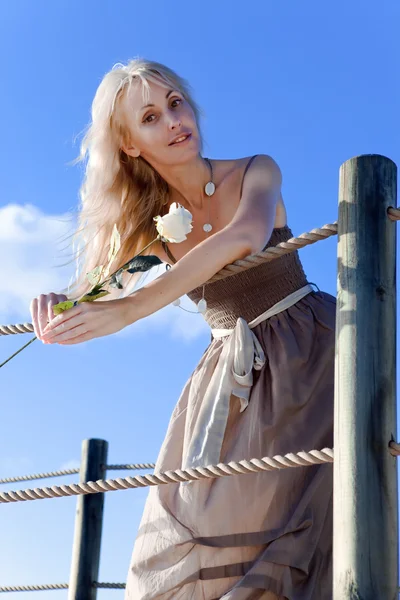 The young beautiful woman with a rose in hands leaned the elbows on ropes on the blue sky a background — Stock Photo, Image