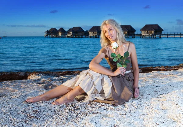 一朵玫瑰的年轻漂亮的女人坐在海边缘的沙滩上 — 图库照片