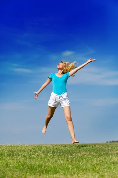 De gelukkige vrouw springt in een zomer groen veld tegen de blauwe hemel — Stockfoto