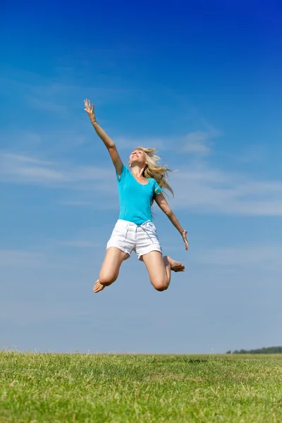 Die glückliche Frau springt in ein sommergrünes Feld gegen den blauen Himmel lizenzfreie Stockbilder