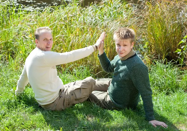 De vader en de zoon zitten op de met gras begroeide kust van het meer — Stockfoto
