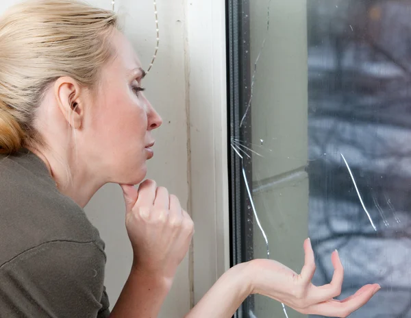 Den sorgliga unga kvinnan nära ett fönster med burst, trasiga glas — Stockfoto