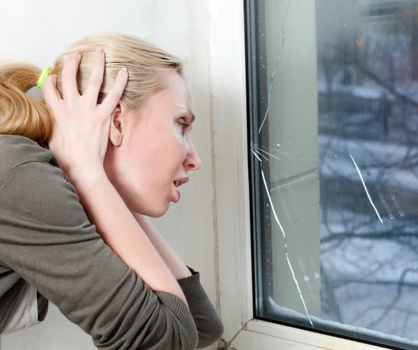 Den sorgliga unga kvinnan nära ett fönster med burst, trasiga glas — Stockfoto