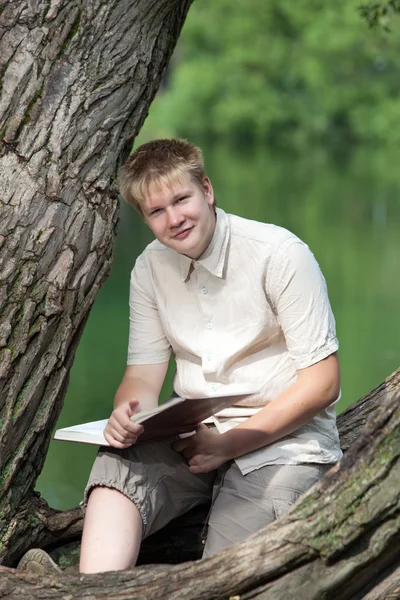 O jovem com o livro no parque na margem do lago — Fotografia de Stock