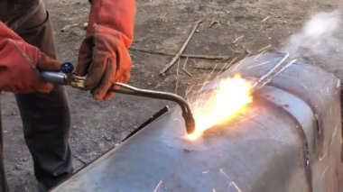 Kaynak makinesinin yangının içinden görüntüsü. Açık havada koruyucu giysiler giyen bir kaynakçı metal boruları asetilen kaynaklama, ateş ve kıvılcımlarla keser..